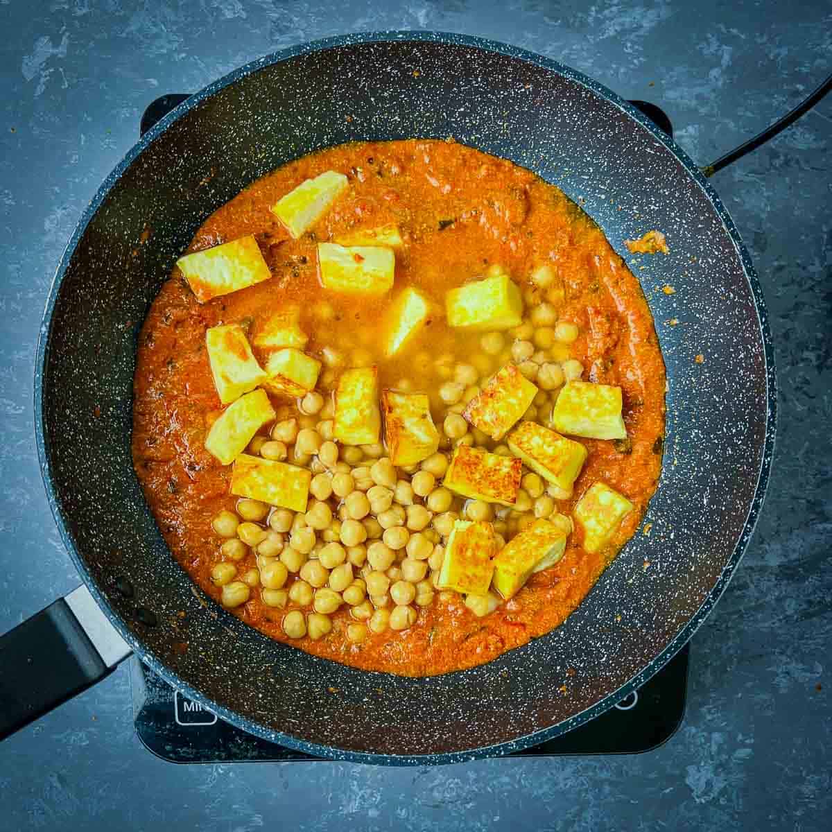 Boiled chickpea and fried paneer added to curry sauce in frying pan.