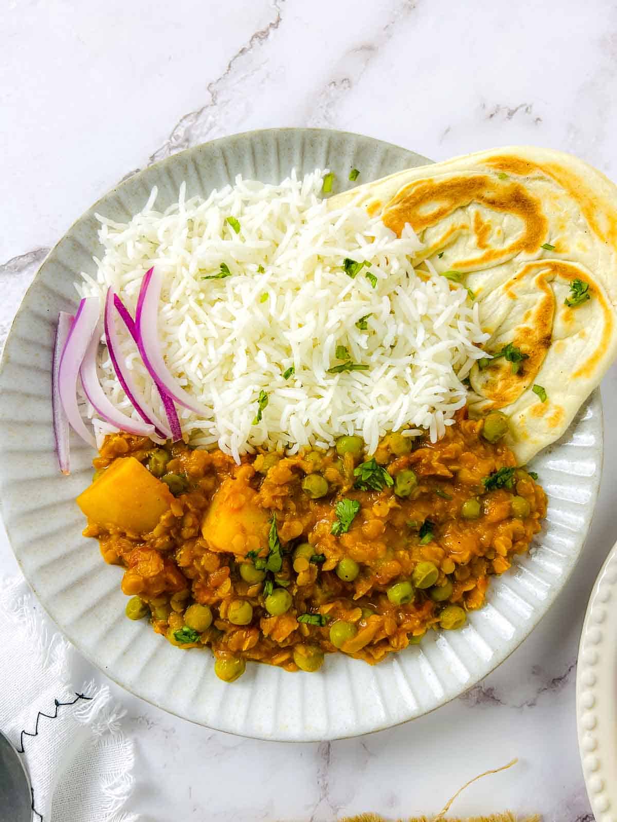 Lentil peas potato curry on a white plate served with rice, naan, and onion slices and garnished with cilantro.
