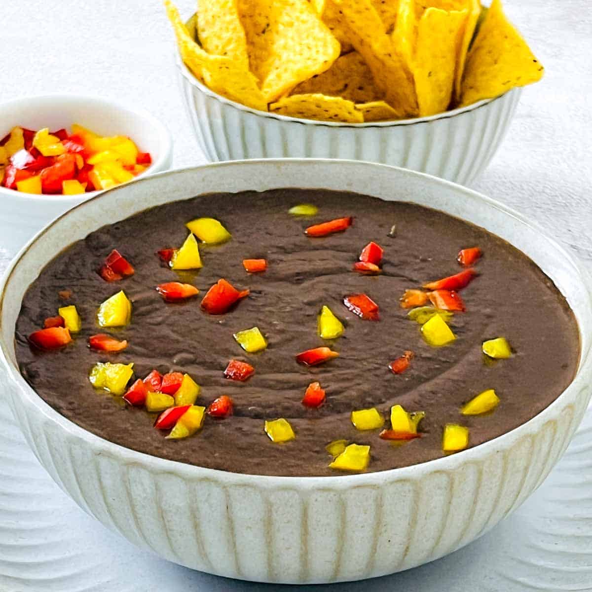 Refried beans in a white bowl with bell peppers and tortilla chips in the background.