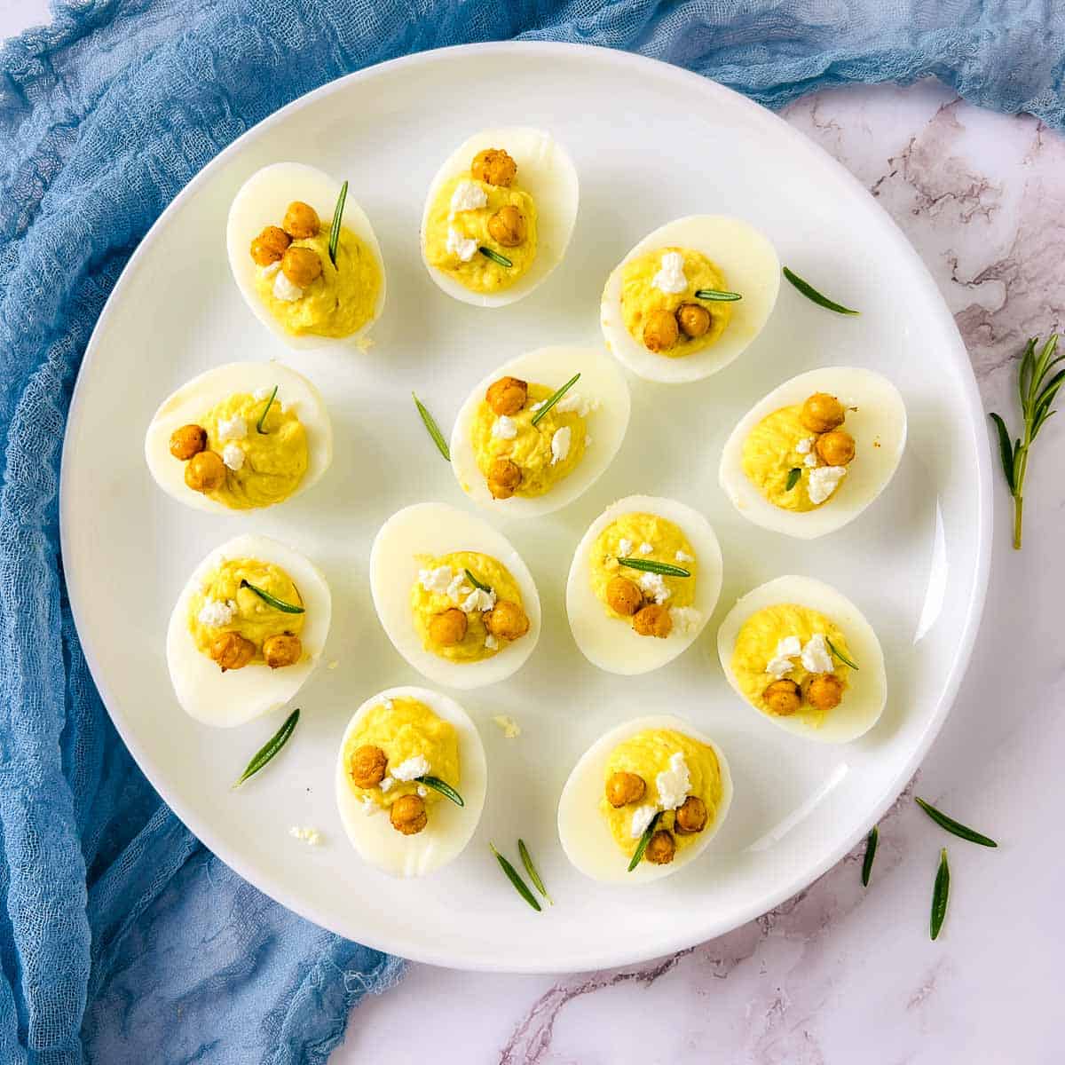 Deviled eggs with roasted chickpeas placed on a white plate with rosemary sprig in the background.
