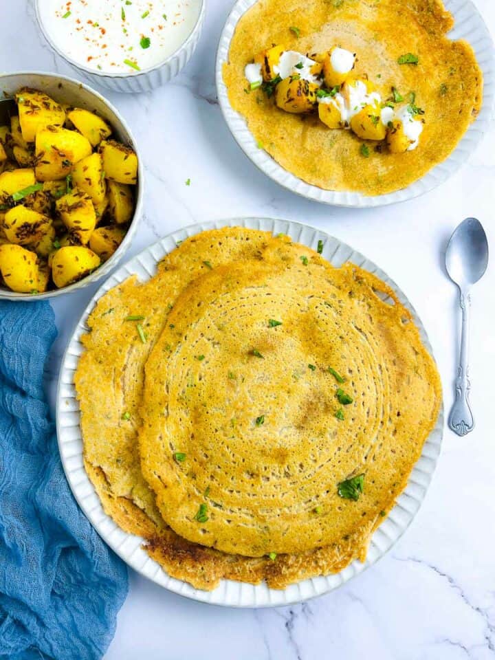 Red lentil pancakes on a white plate with potatoes and raita in the background.