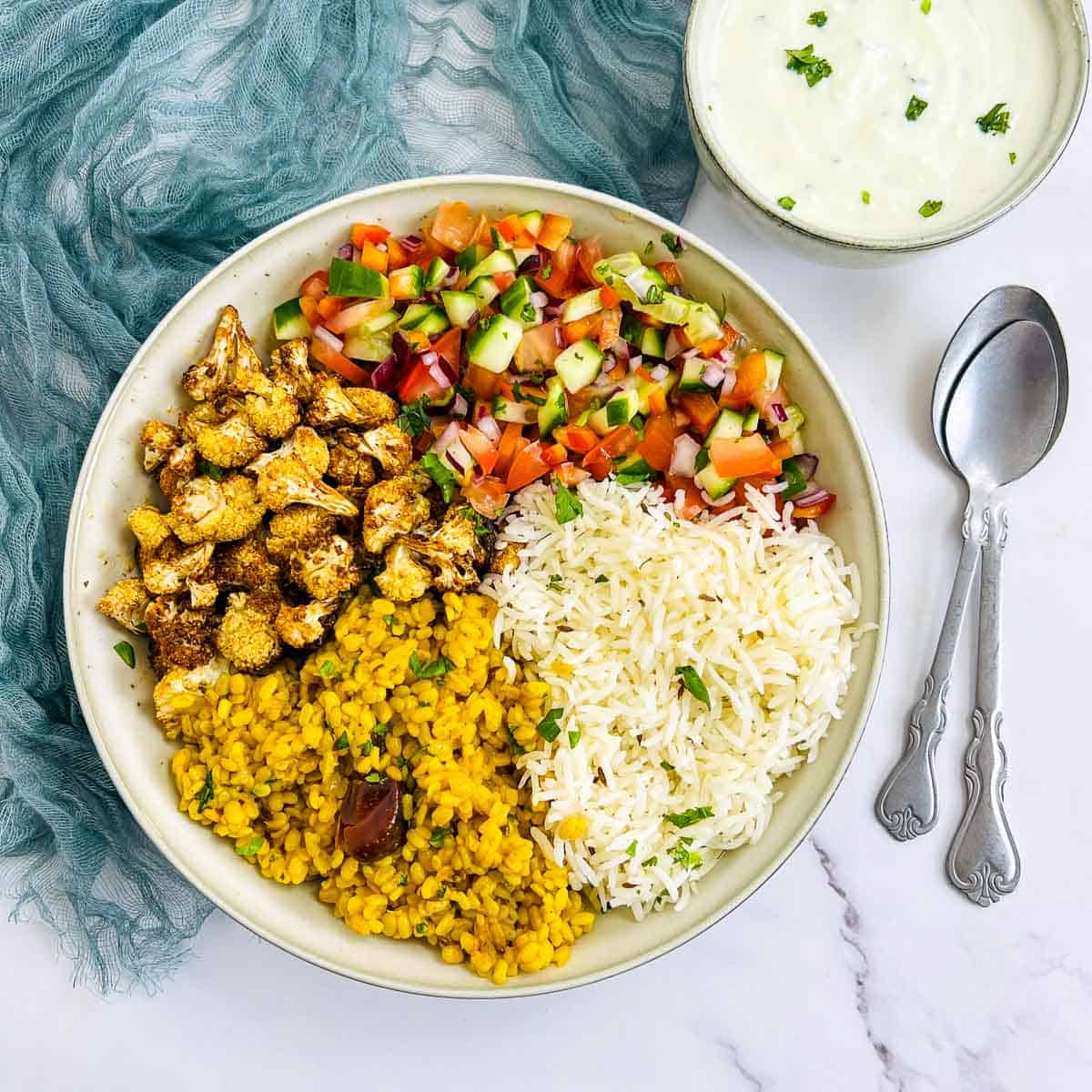 Yellow lentil Indian Buddha bowl and raita bowl on a white surface with spoons in the background.