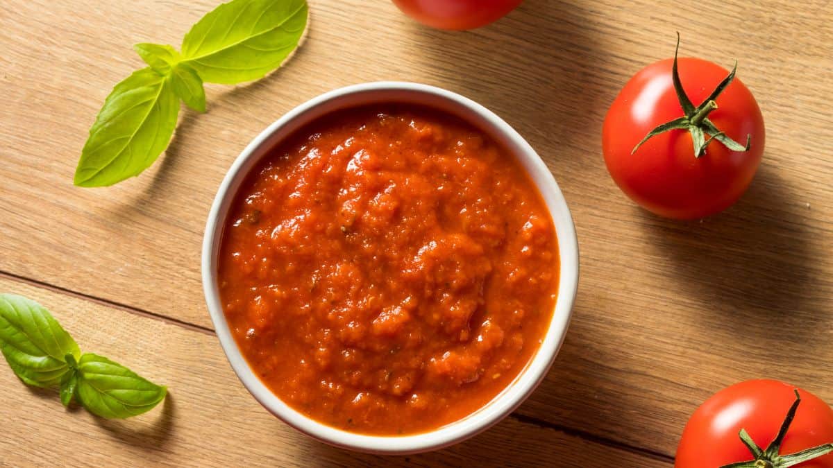 Pasta sauce in a bowl with tomatoes and basil in the background.