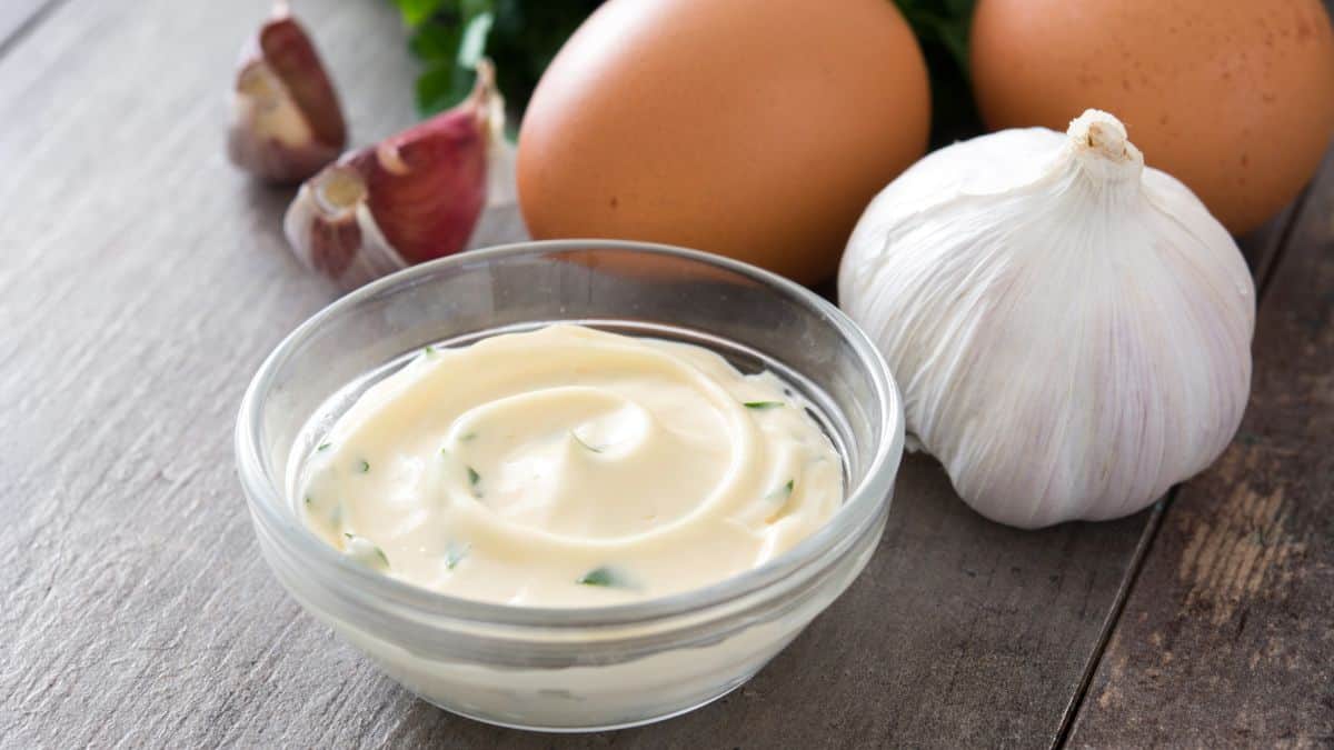 Aioli in a small bowl with garlic and eggs in the background.