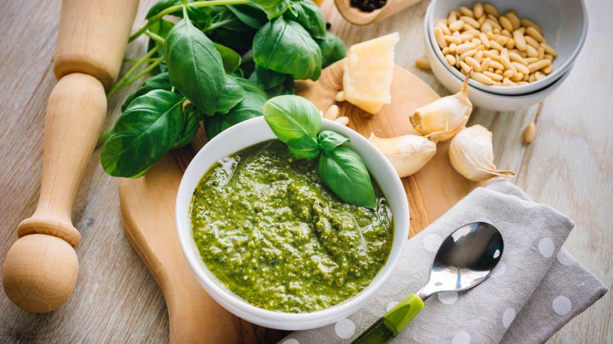 Pesto sauce in a white bowl with ingredients in the background.