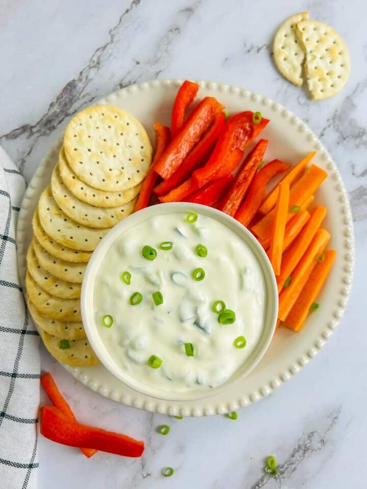 Green onion dip in a white bowl served with crackers and veggie sticks.