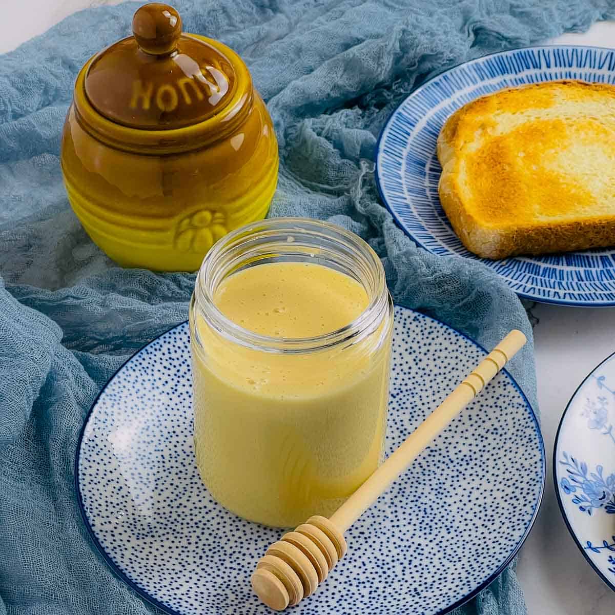 Whipped honey jar placed on a plate with honey drizzle spoon and toasts in the background.