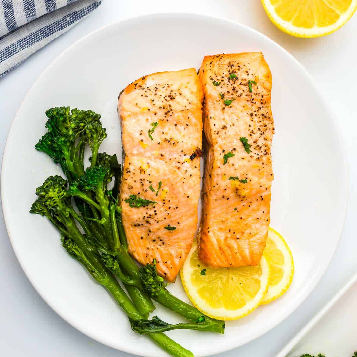 Air fryer salmon on a white round plate served with lemon wedges and broccolini.