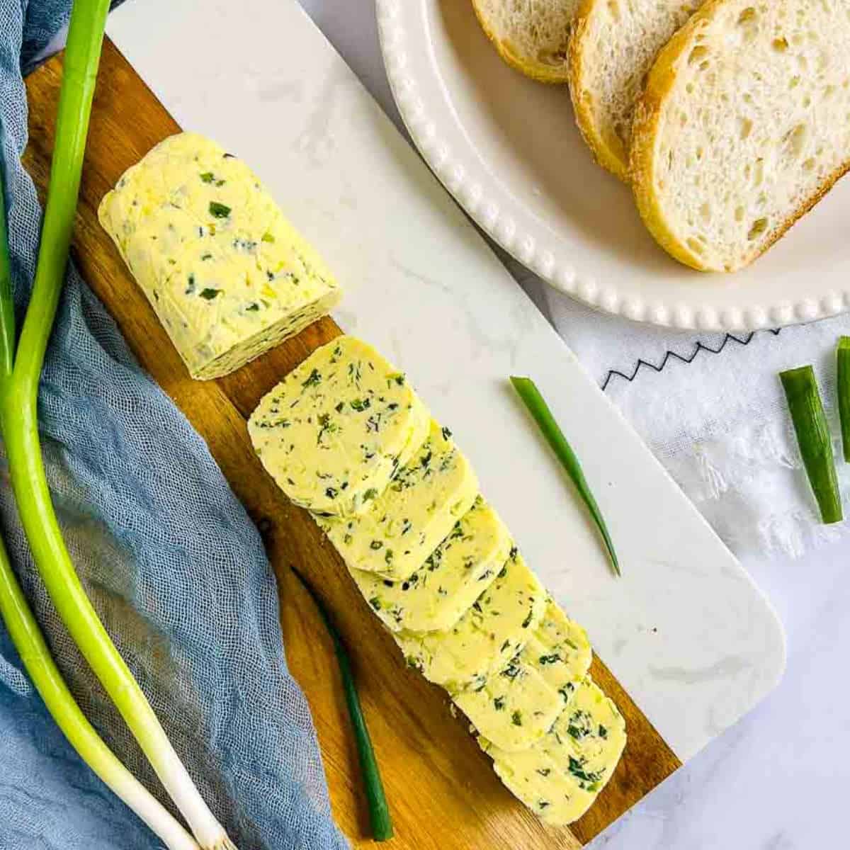 Log of scallion butter on a wooden board with bread slices in the background.