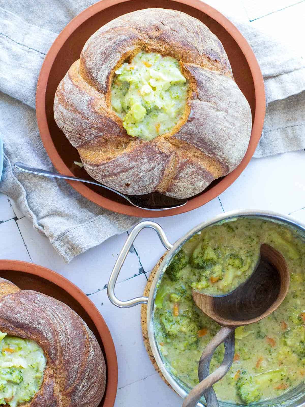 Bread bowls filled with broccoli cheddar soup with soup pot in the background.