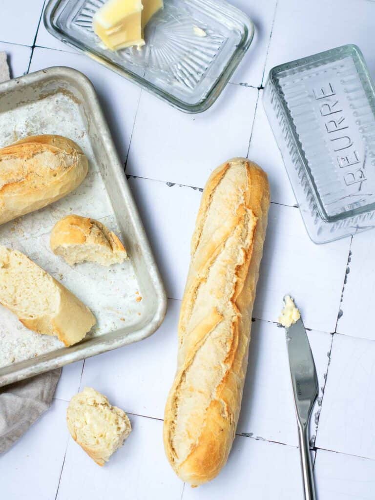 Baguette on a white surface with butter and knife. It also has a sliced baguette placed on a baking sheet.