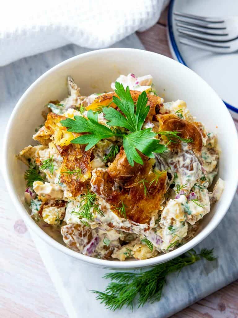 Smashed potato salad garnished with parsley and dill with plates and fork in the background.