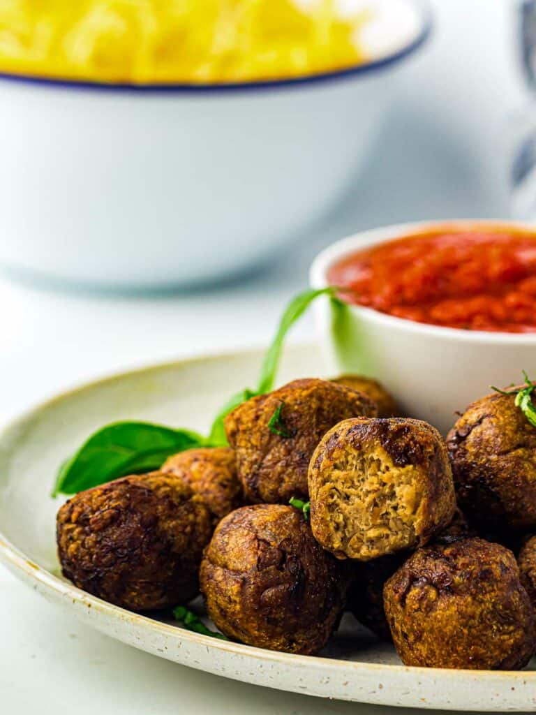 Cooked meatballs served with sauce along with pasta in the background.