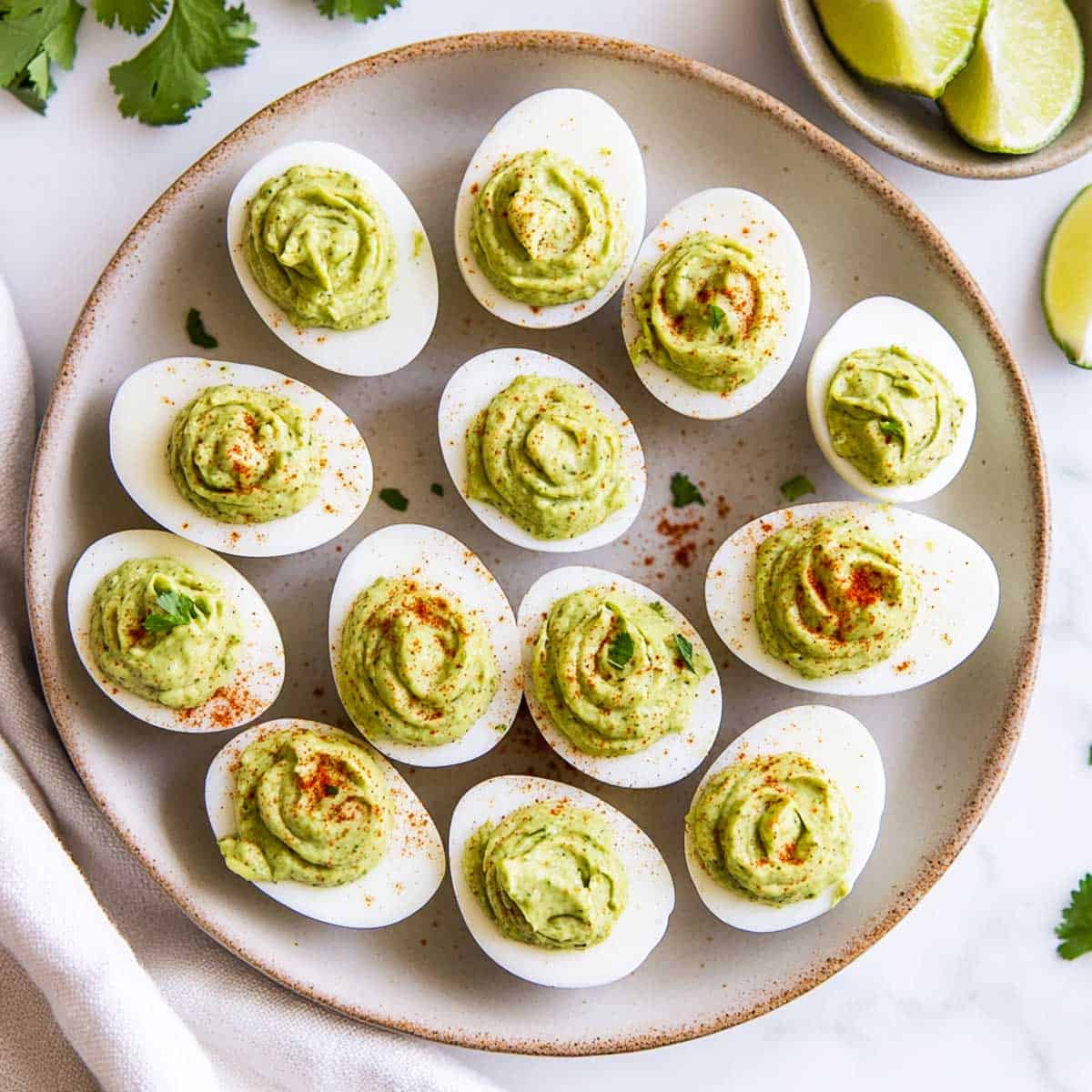 Avocado deviled eggs on a brown plate with cilantro and lime in the background.