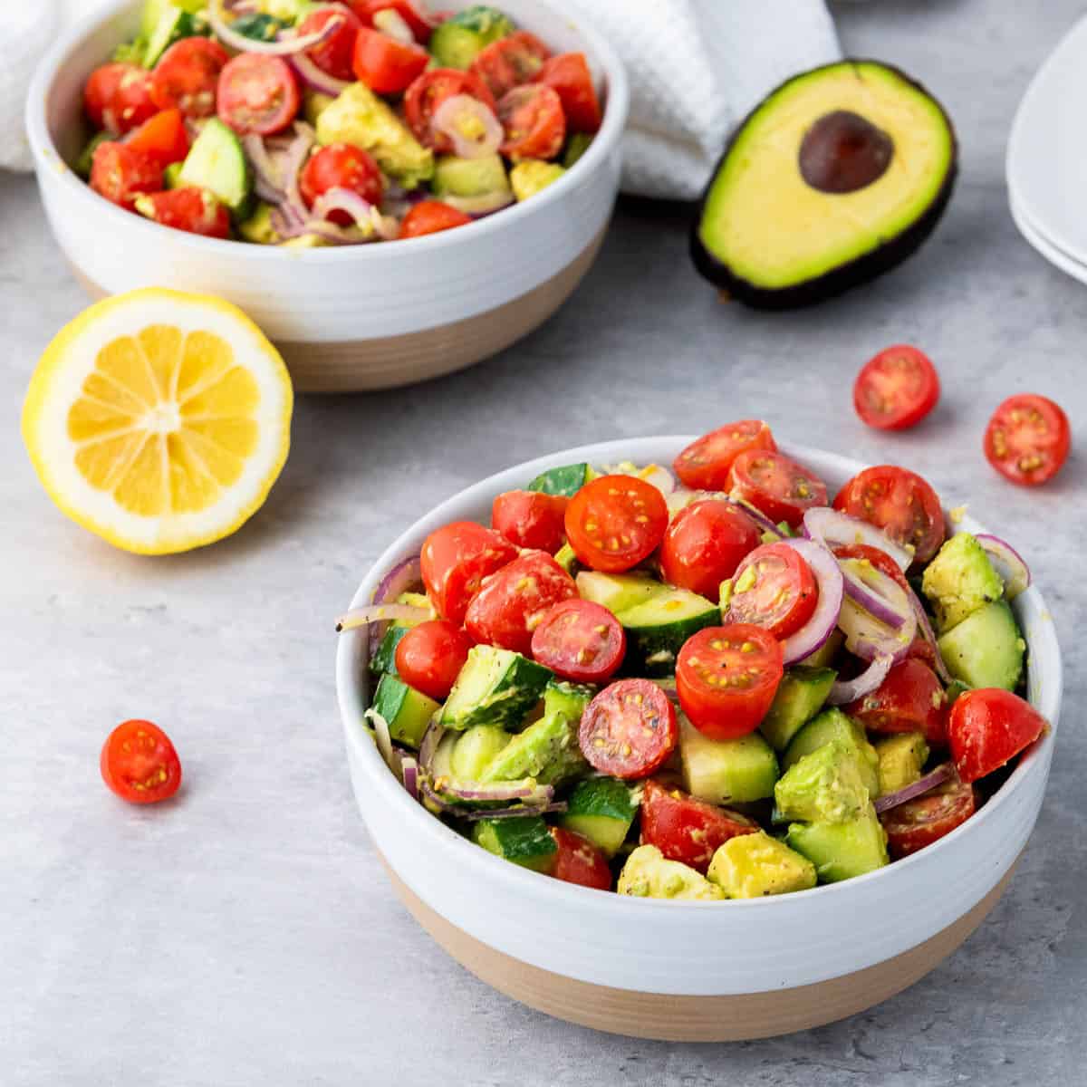 Side view of two bowls of avocado salad placed on a grey surface with lemon, whole avocado, and cherry tomatoes in the background.