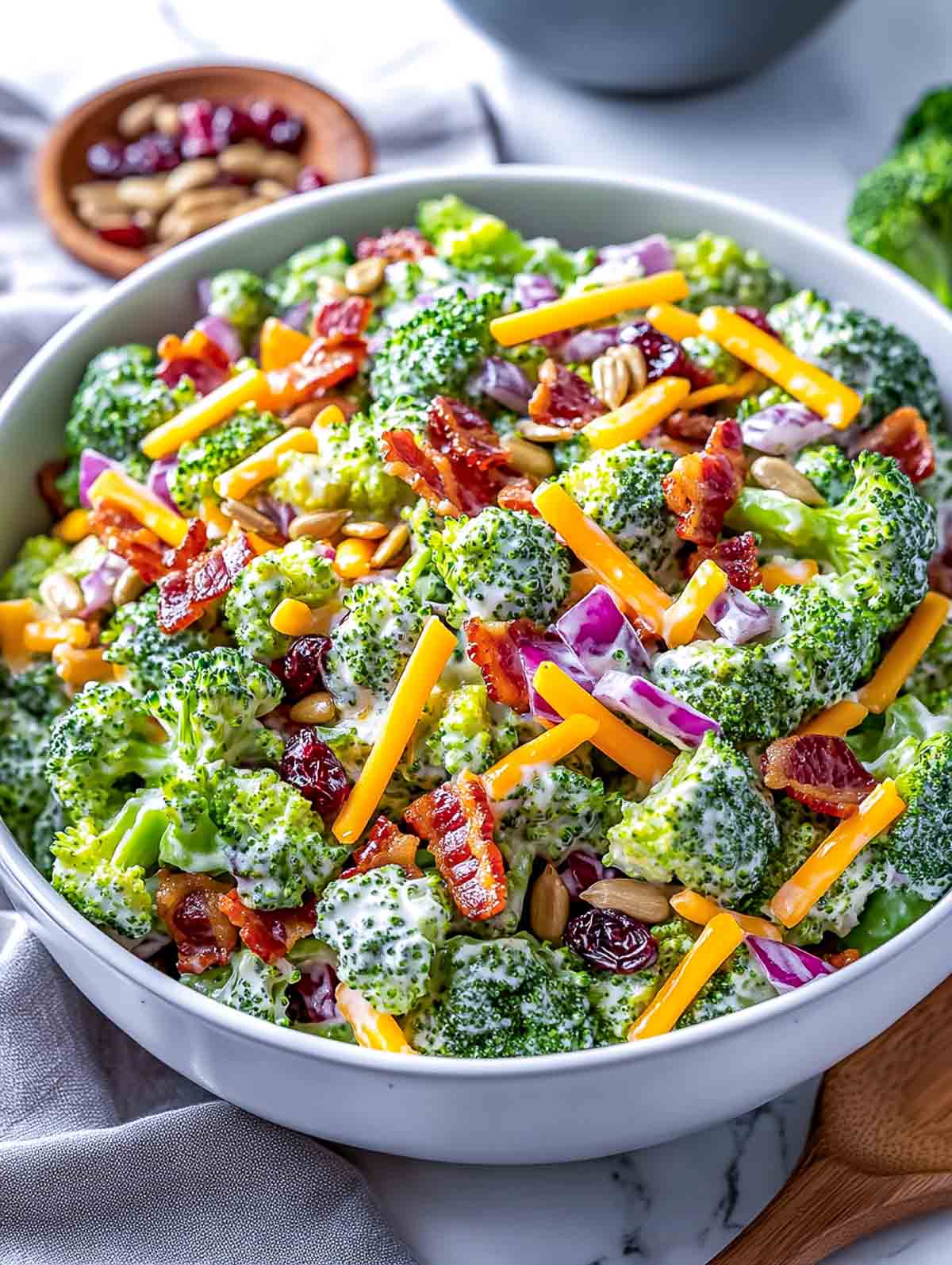 Side view of broccoli salad in a wide serving bowl with dried cranberries and almonds in the background.