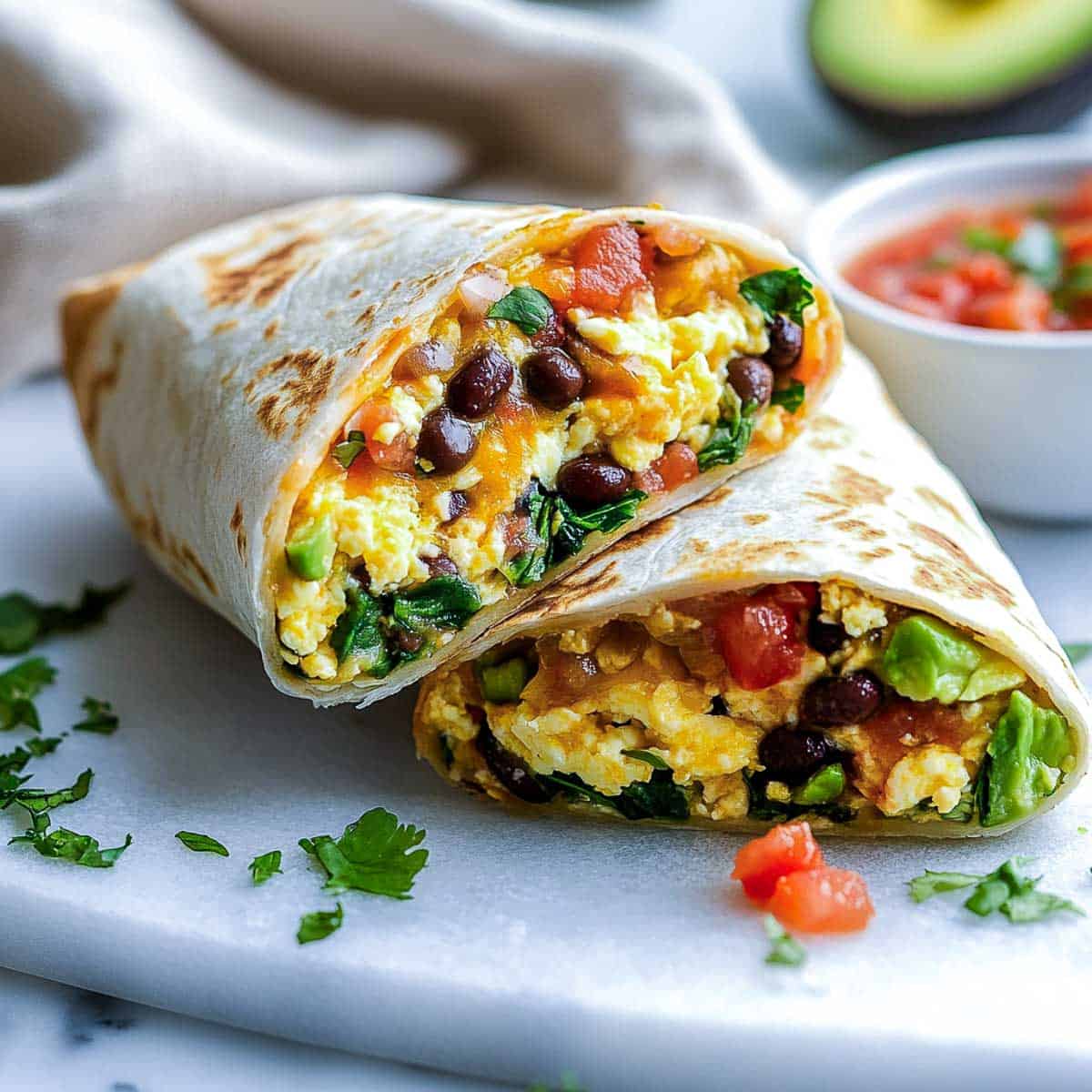 Breakfast burrito cut and placed on a plate with salsa and avocado in the background.