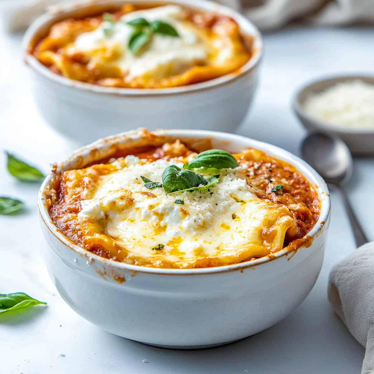 Two lasagna bowls with spoon and a small bowl of cheese in the background.