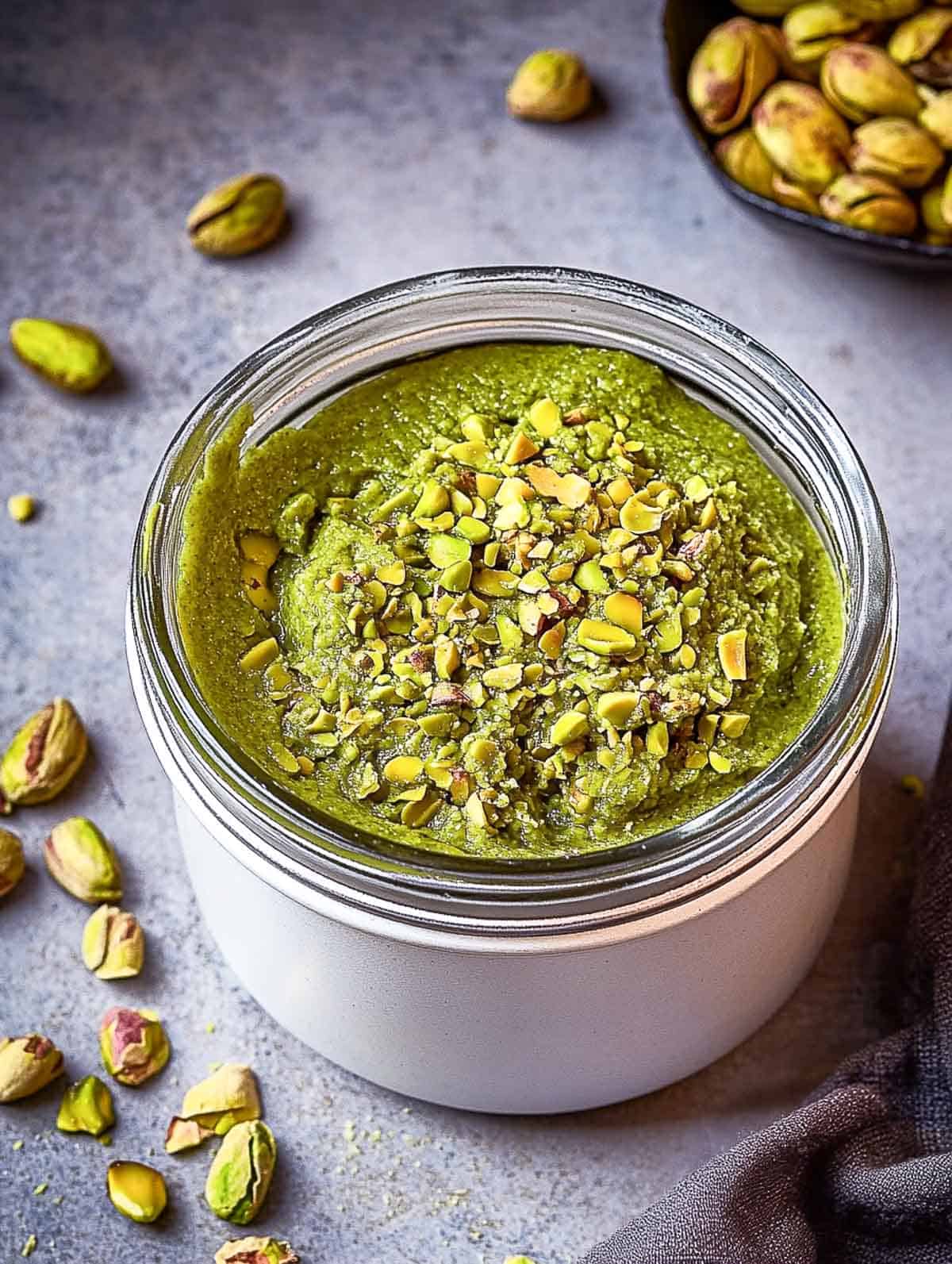 A jar of pistachio paste on a grey surface with pistachios in the background.