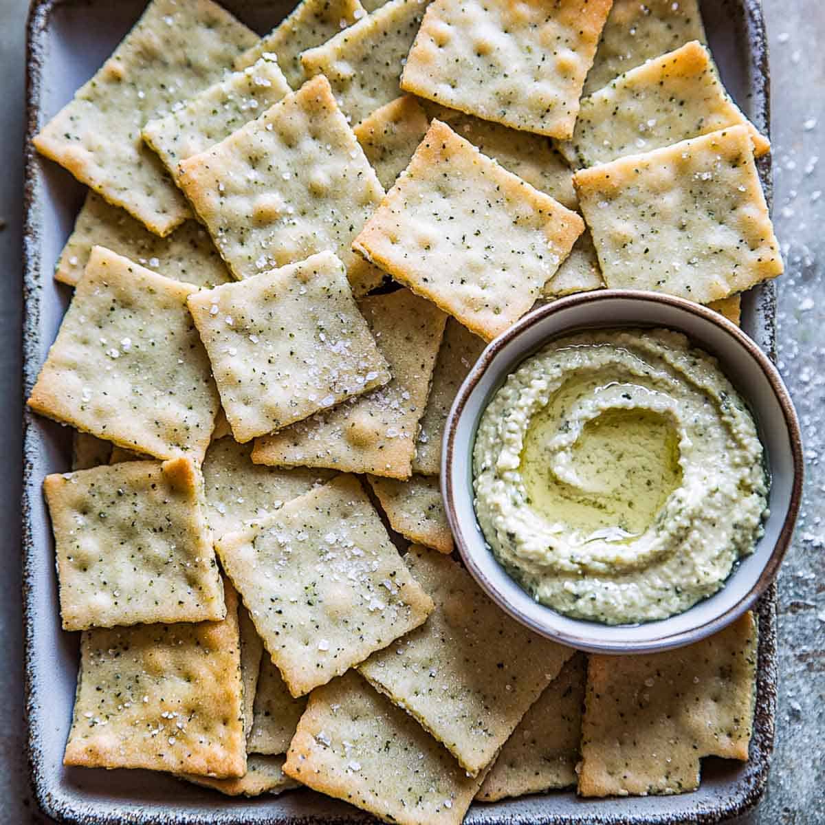 Crispy crackers on a baking tray with a bowl of herbed hummus in the side.