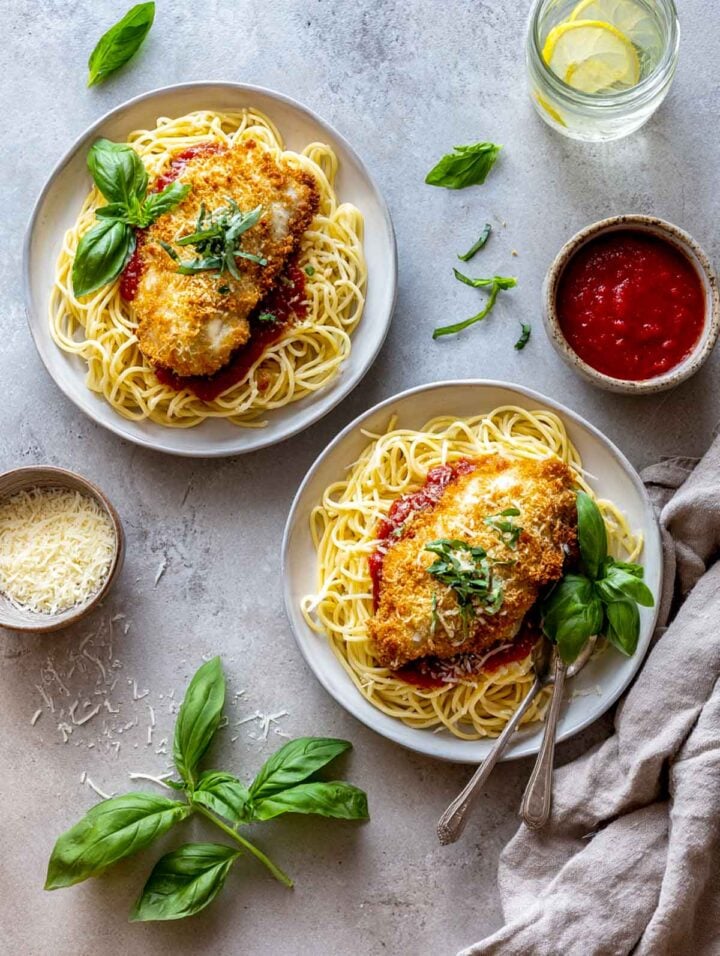 Top view of Chicken Parmesan with spaghetti on two plates, surrounded by fresh basil, shredded cheese, and a napkin.