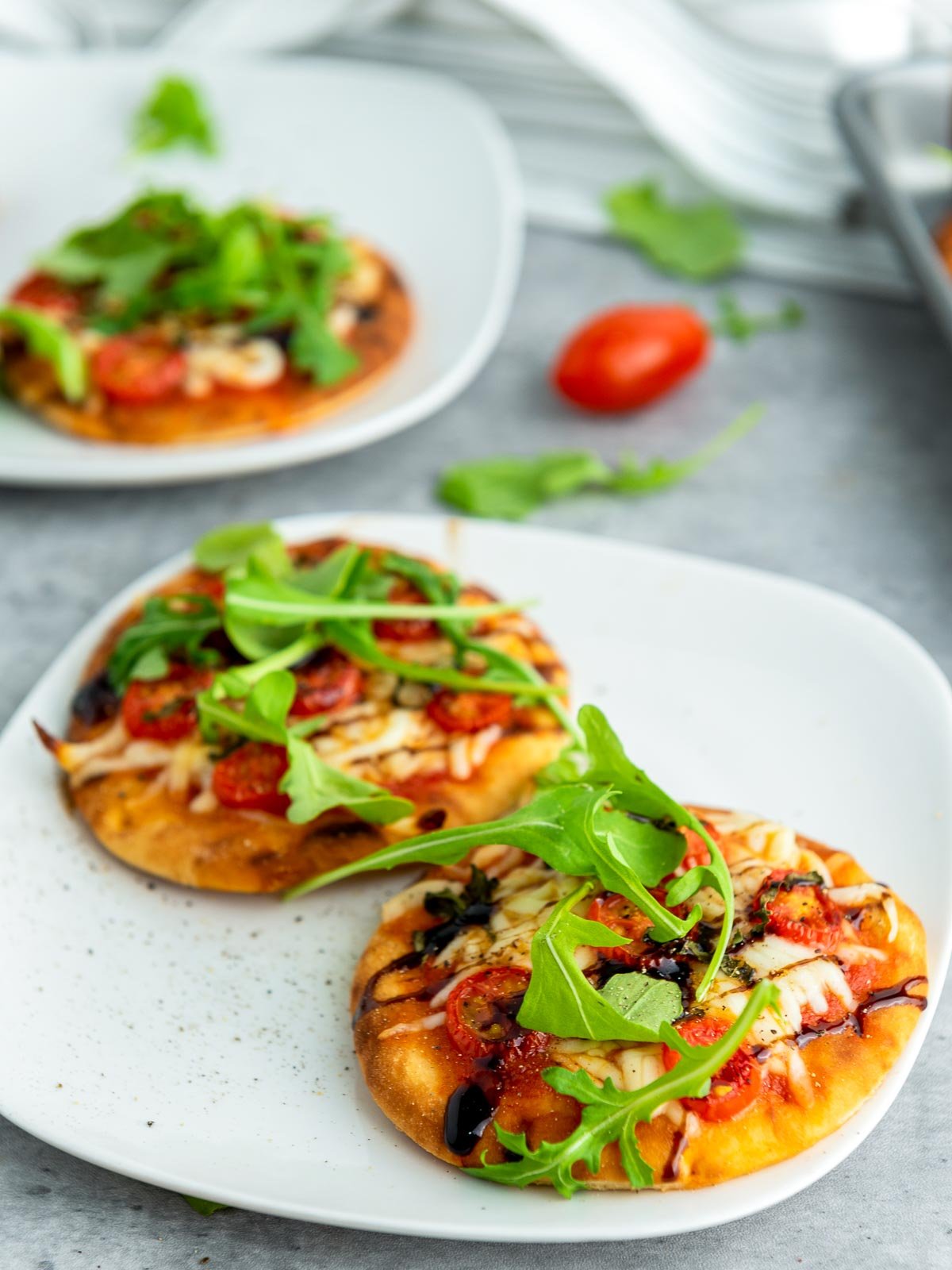 Two mini Caprese pizzas on a plate, garnished with fresh arugula and surrounded by tomatoes and greens on a gray surface.