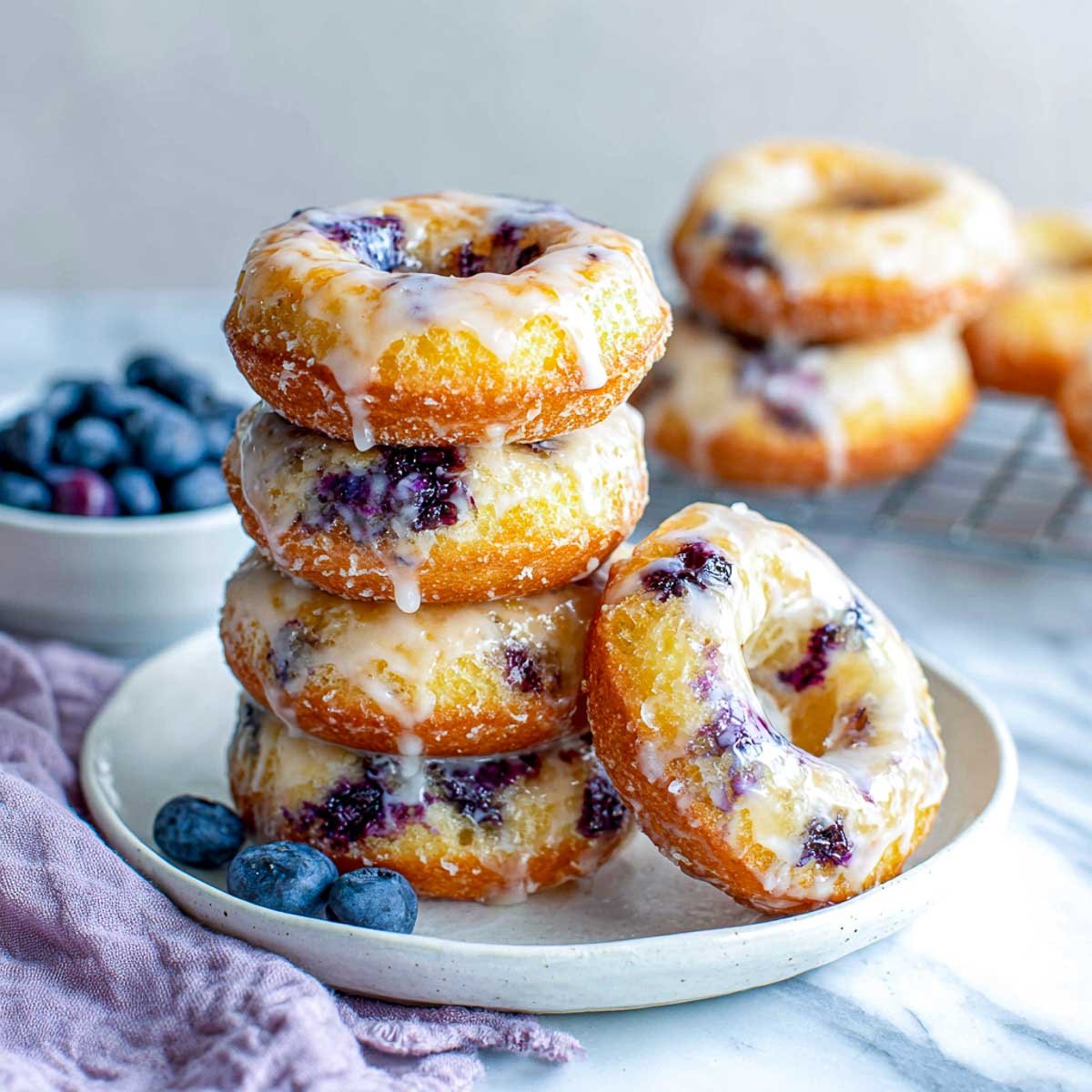 Air fryer blueberry donuts stacked on a white plate with glaze dripping, fresh blueberries on the side.