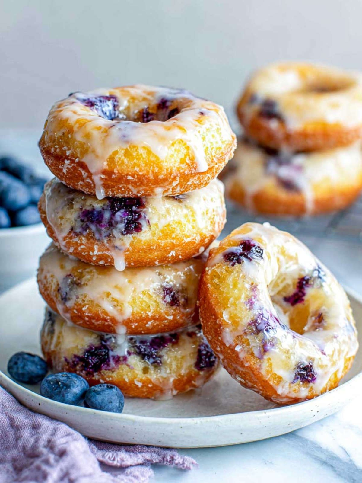 Close-up of stacked air fryer blueberry donuts on a white plate with visible glaze and fresh blueberries.