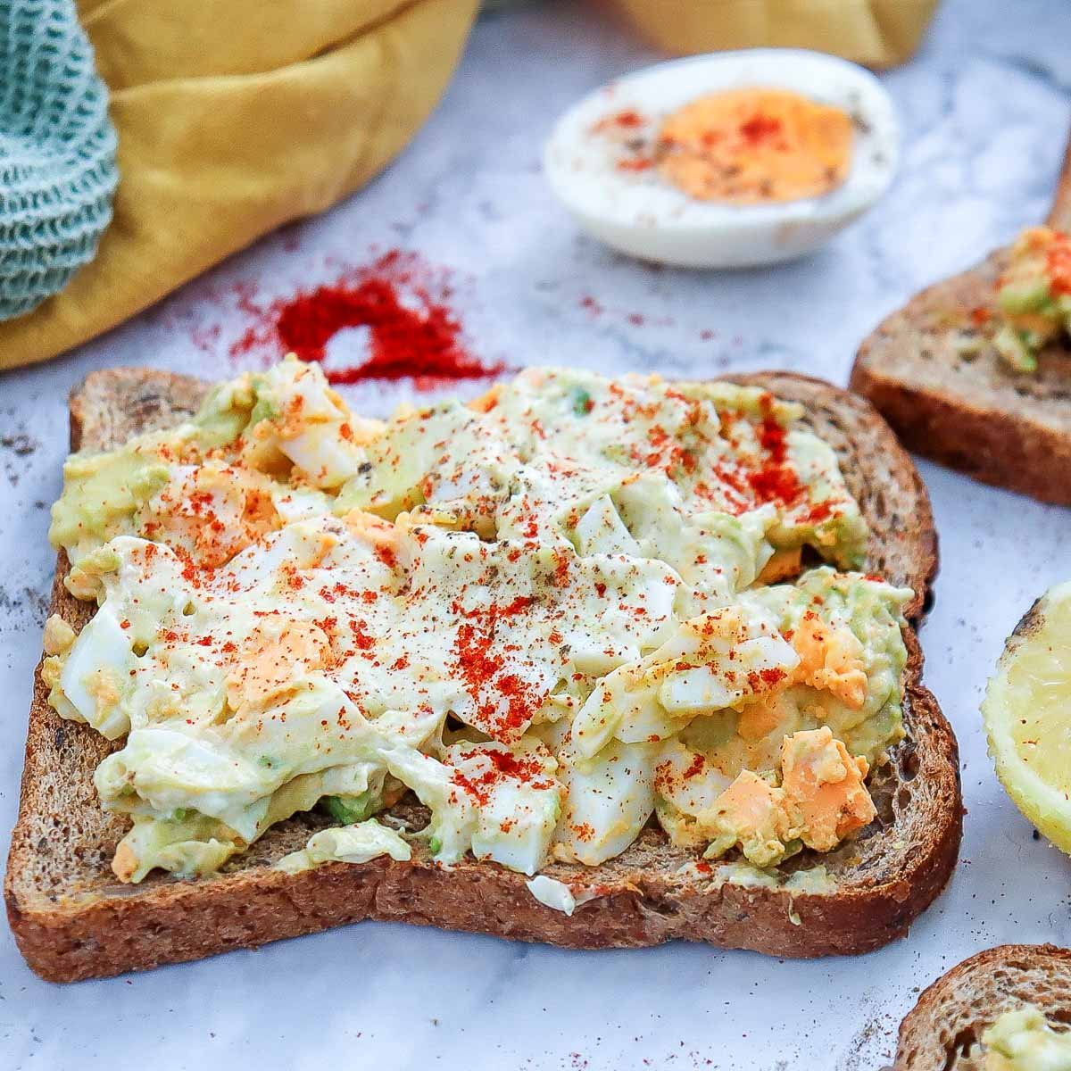 Angled shot of avocado egg salad toast with creamy topping, surrounded by paprika, lemon, and a hard-boiled egg half.