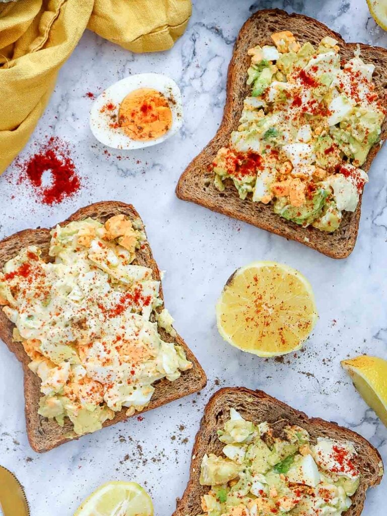 Top-down view of multiple slices of avocado egg salad toast on marble background with lemon halves, paprika, and a hard-boiled egg.