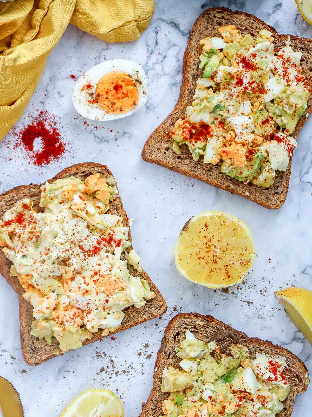 Top-down view of multiple slices of avocado egg salad toast on marble background with lemon halves, paprika, and a hard-boiled egg.