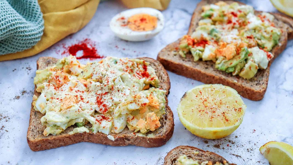 Side view of avocado egg salad toast surrounded by lemon, paprika, and bread slices, with a rustic presentation.