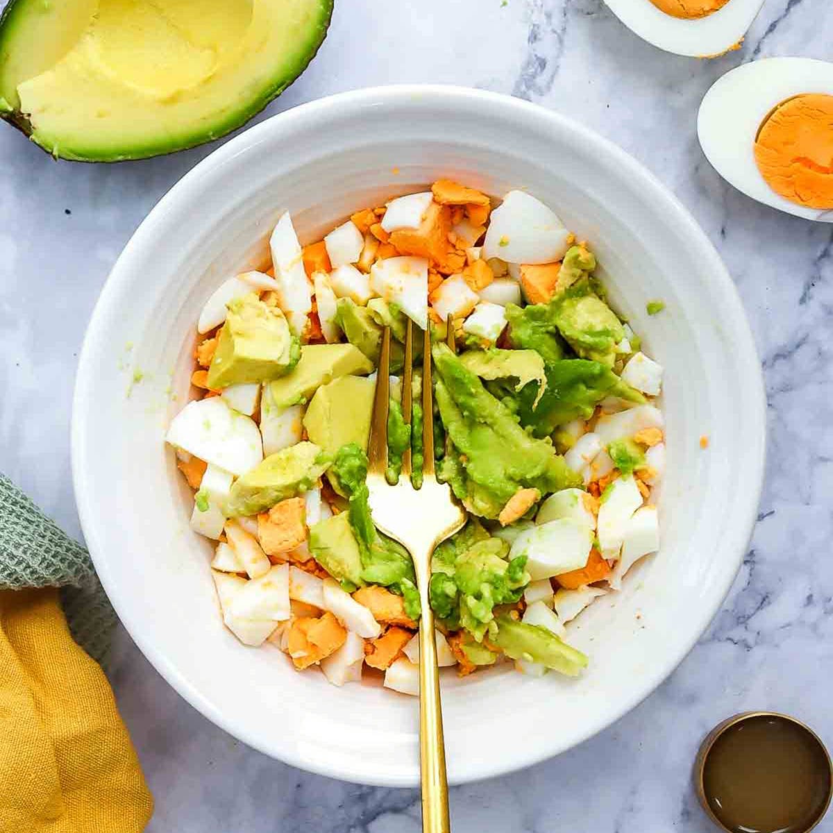 Chopped boiled eggs and avocado chunks in a white bowl with a fork, ready to be mashed together.