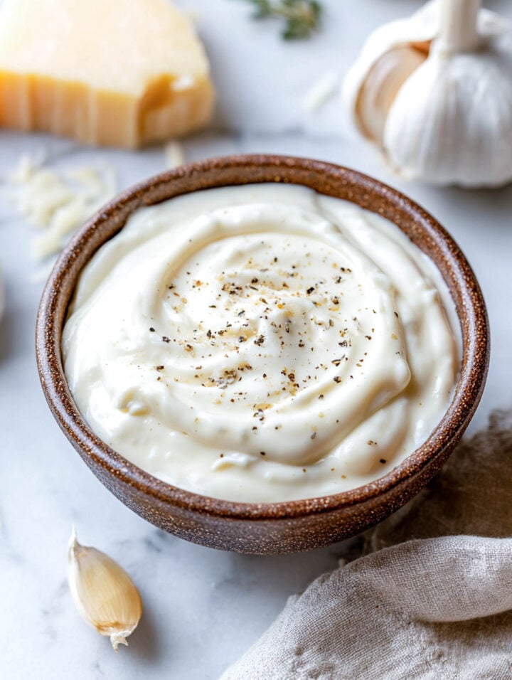 Creamy cottage cheese Alfredo sauce swirled in a brown ceramic bowl with black pepper, set against garlic and Parmesan in the background.