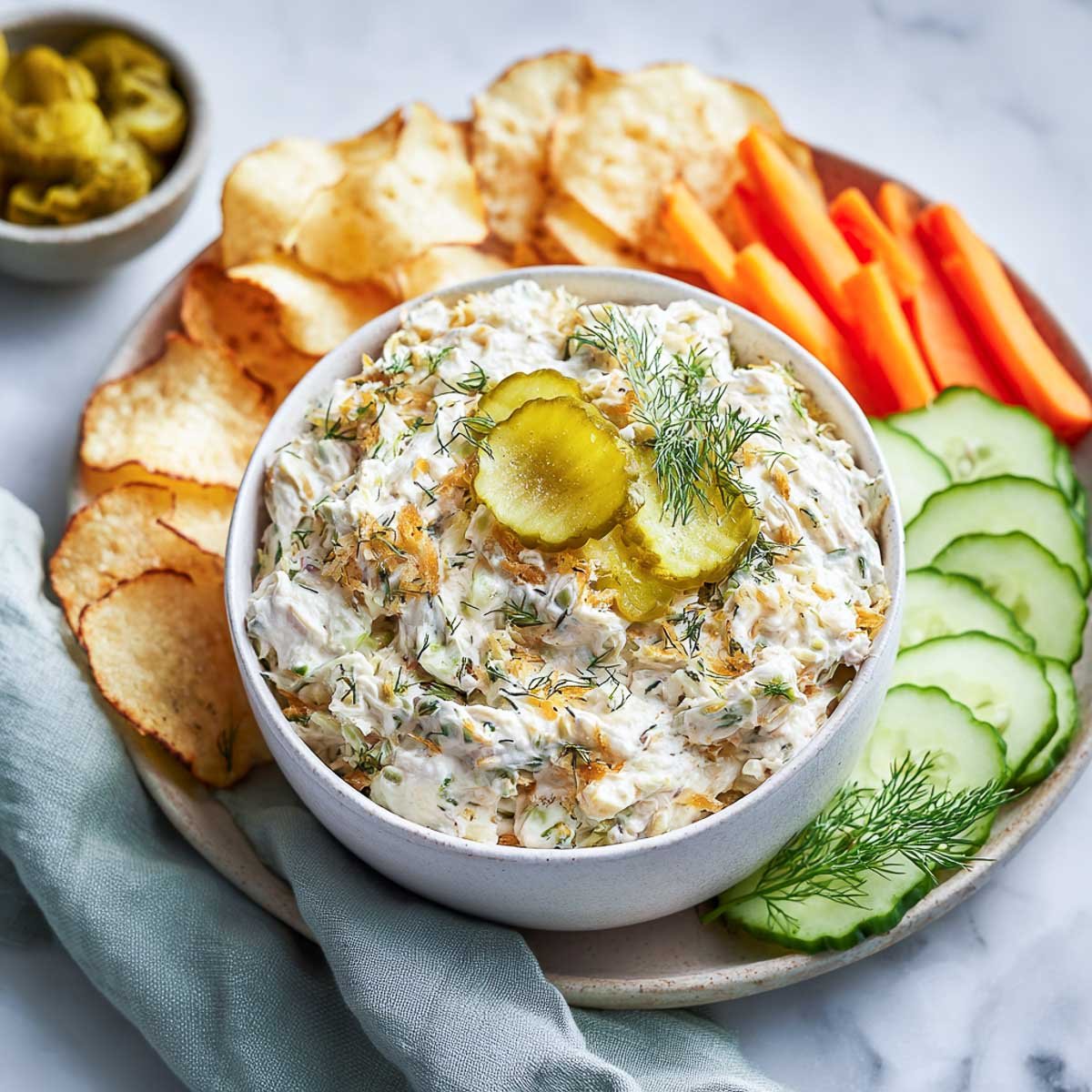 Chunky dill pickle dip with herbs and pickles, served with chips, carrots, and cucumbers.