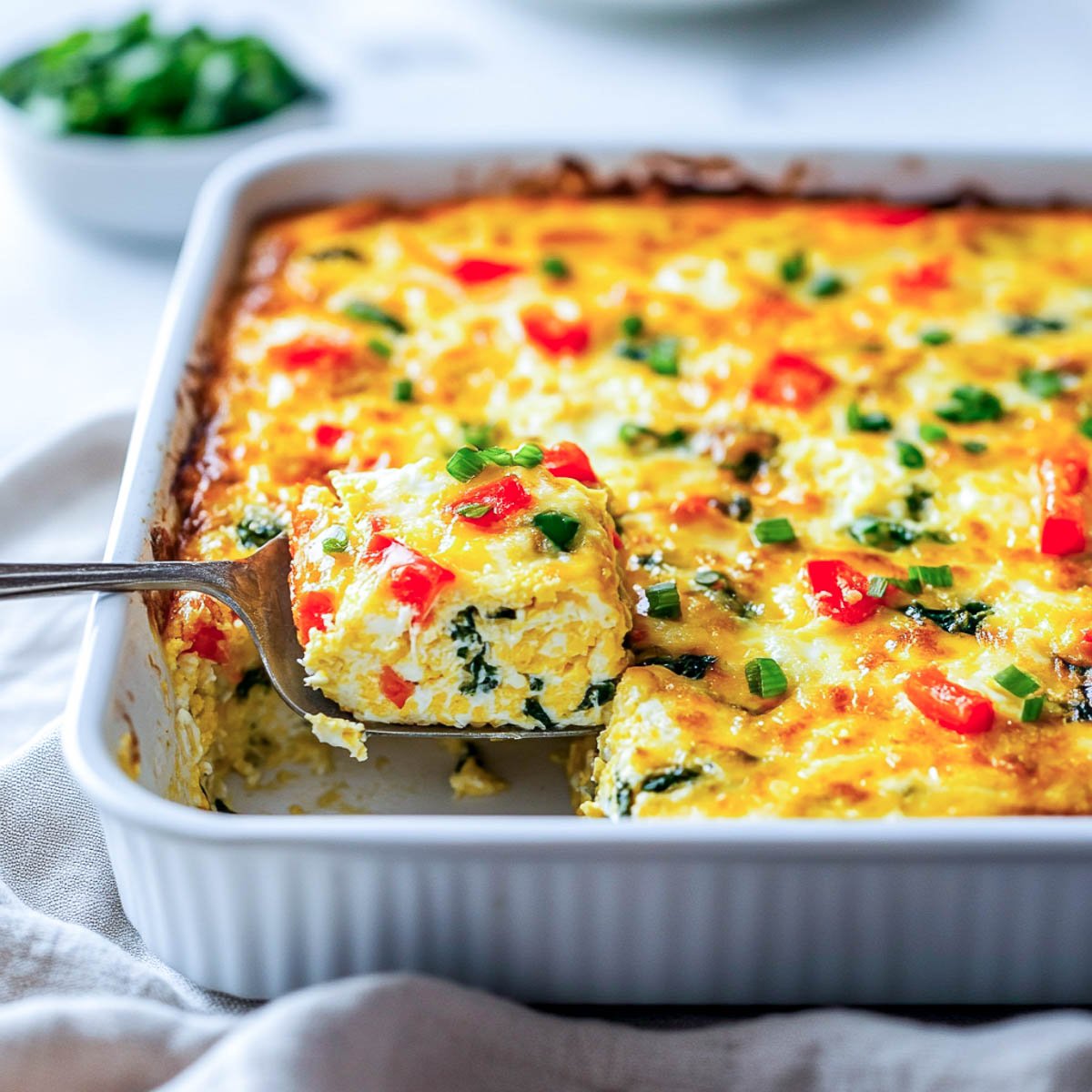 Close-up of a cottage cheese egg casserole in a white baking dish, with a slice being lifted to show the fluffy texture, colorful bell peppers, spinach, and melted cheese on top.