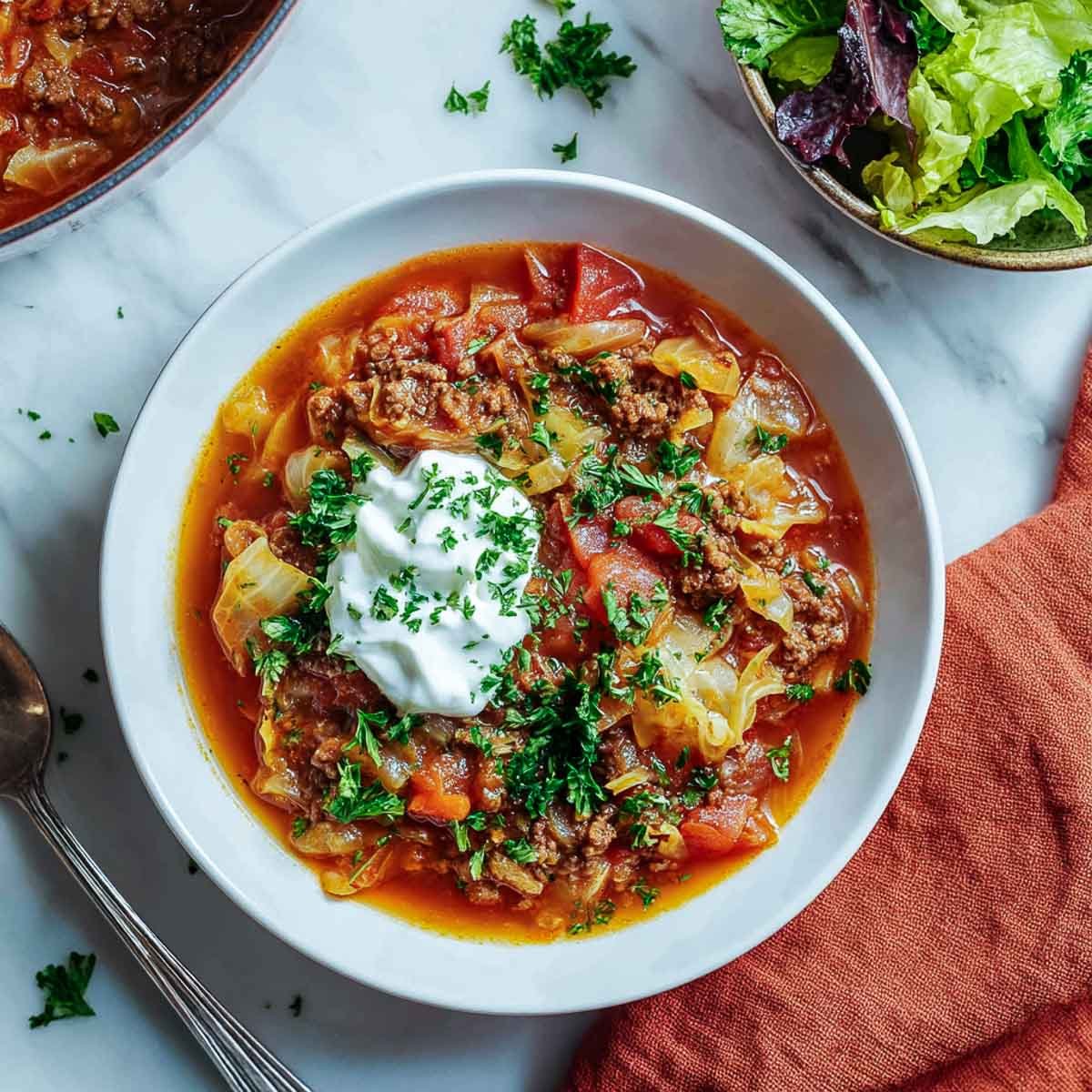 Top-down view of cabbage roll soup in a white bowl with salad and sour cream.