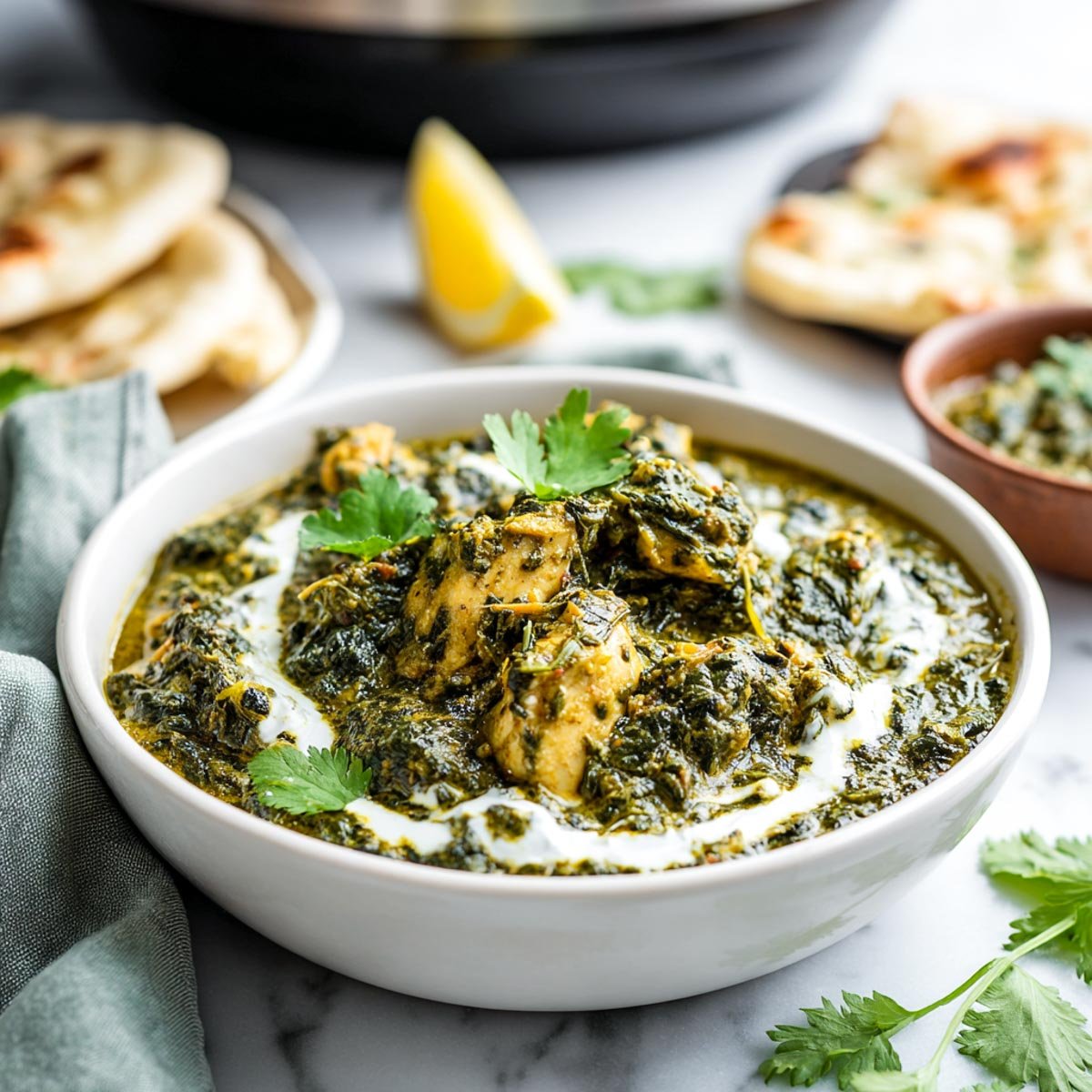 Slightly angled view of saag chicken in a white bowl, showing the rustic spinach texture and chunks of chicken, garnished with yogurt and cilantro. Naan and a lemon wedge are in the background, with a sage green napkin on white marble.