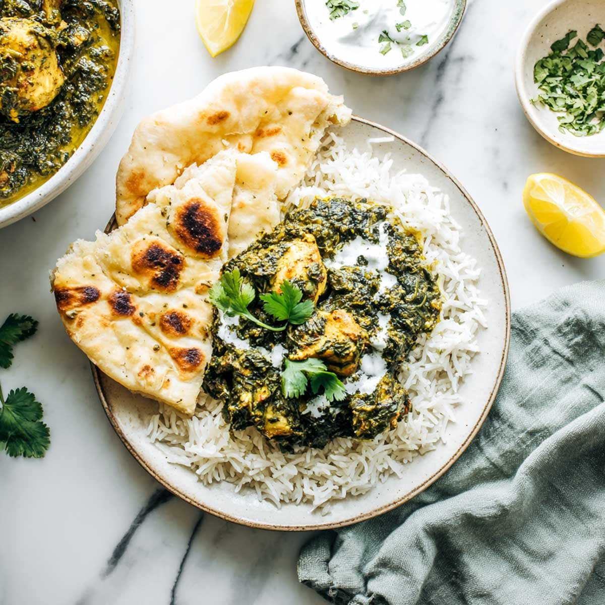Top-down image of Crockpot Saag Chicken served over basmati rice with naan on the side, garnished with yogurt and cilantro. Background includes a raita bowl, lemon wedge, and serving bowl on white marble with a sage green napkin.