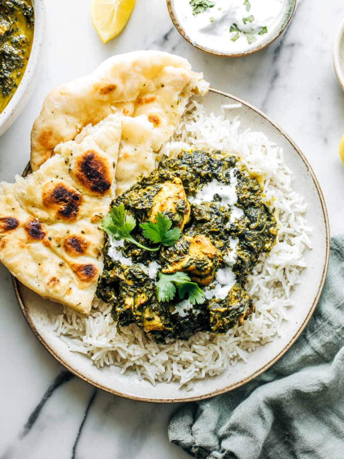 Close-up top-down view of saag chicken on a plate of basmati rice with naan, showing the spinach curry’s texture and yogurt garnish. Accompanied by a green linen napkin, raita, and lemon wedge on a white marble surface.