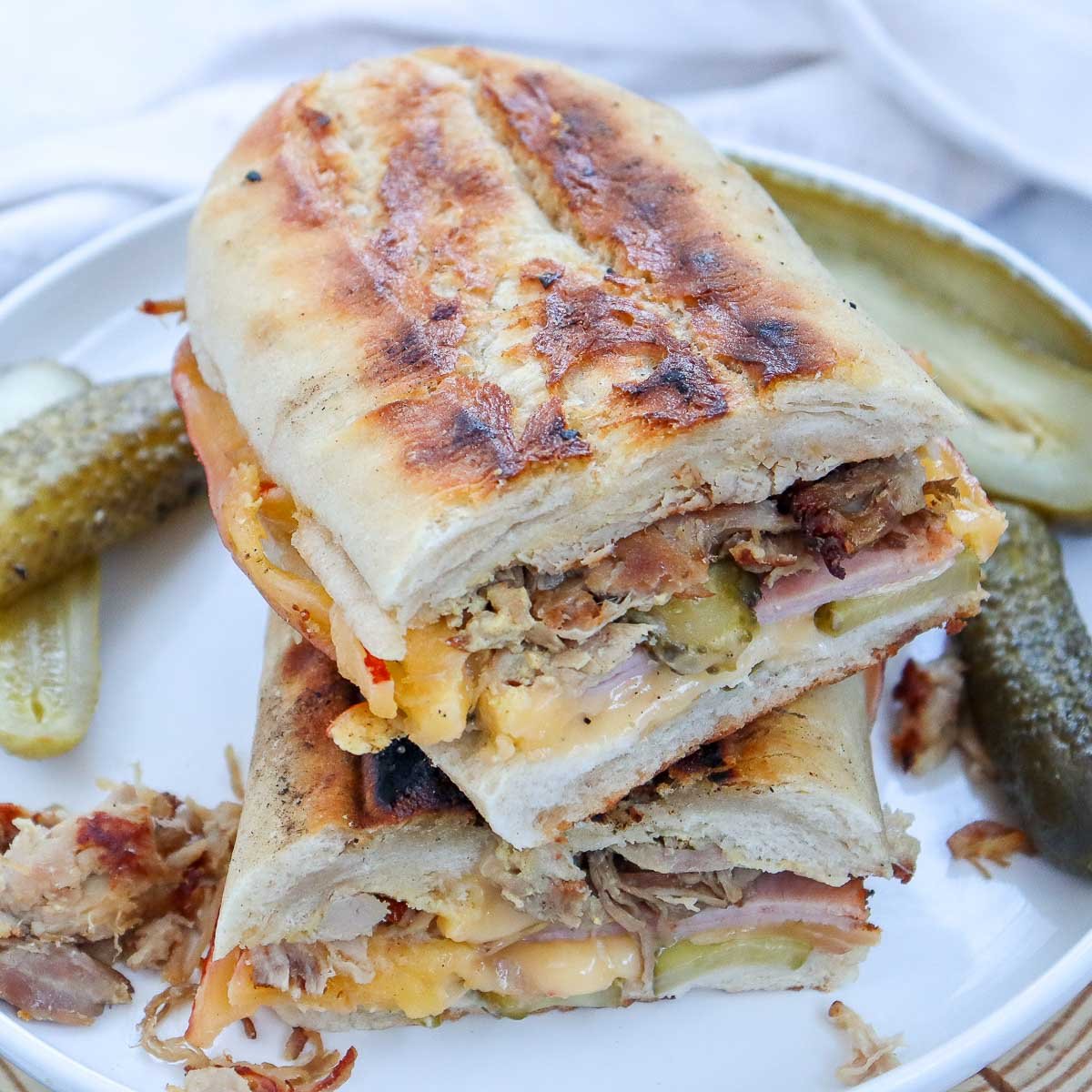 Grilled Cuban sandwich stacked on a white plate with pickles on the side.