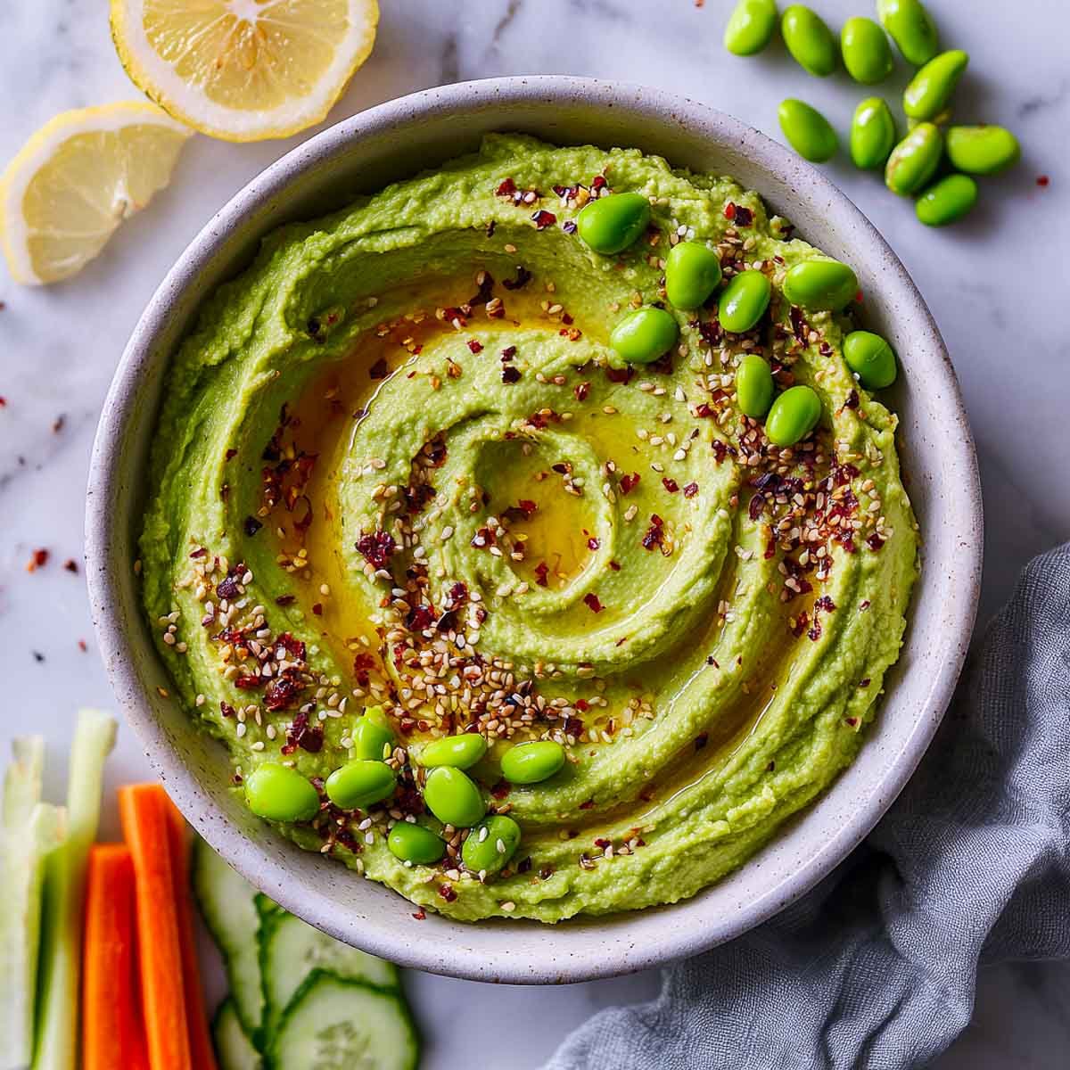 Top-down view of green edamame hummus with sesame, oil, and veggie sticks.