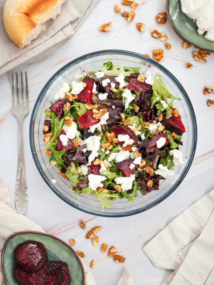 Top-down shot of beet salad with goat cheese and walnuts surrounded by ingredients on a marble surface.