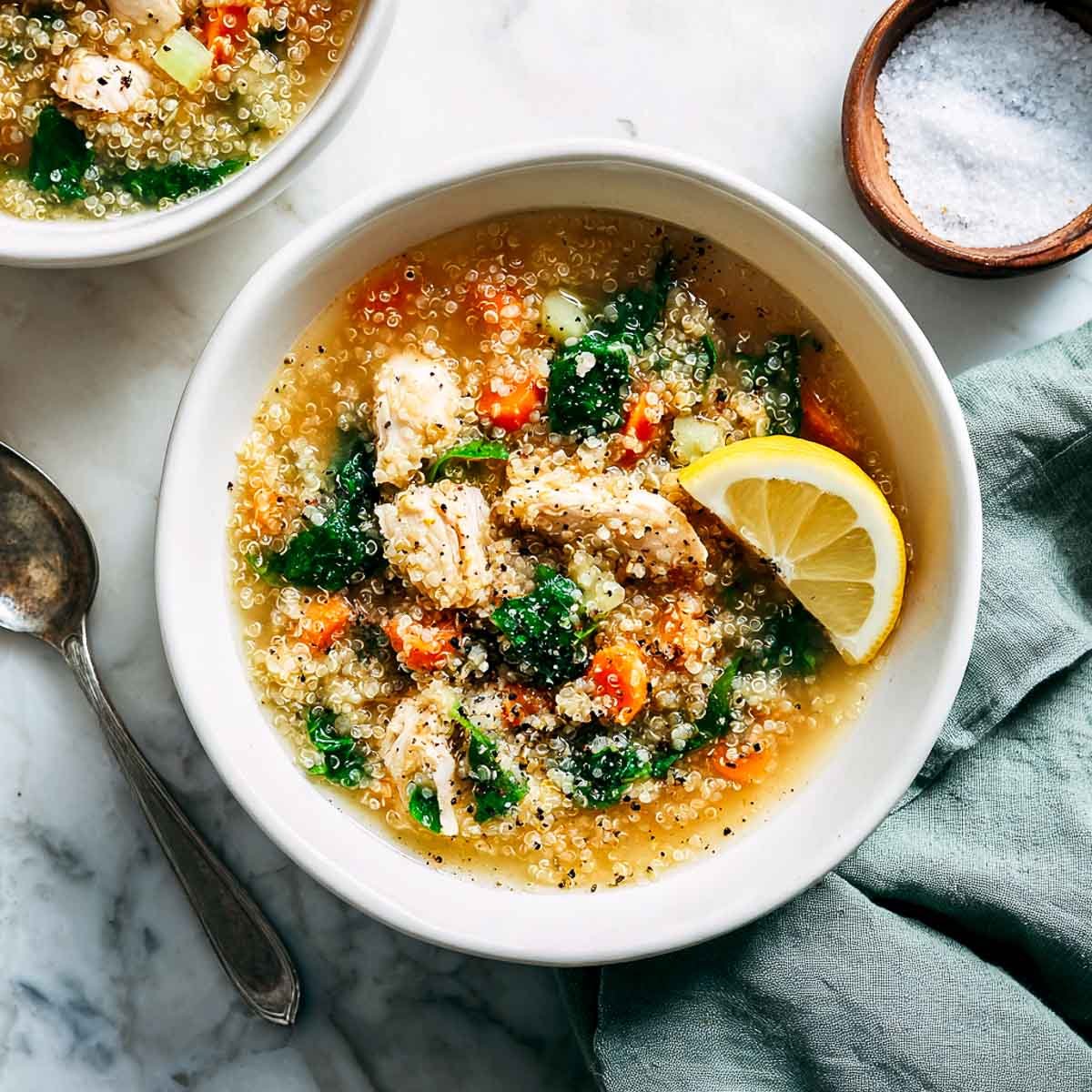 Brothy chicken quinoa soup with spinach, carrots, and lemon in a white bowl.