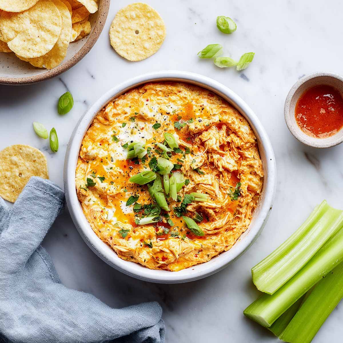 Buffalo chicken dip in white bowl topped with scallions, celery, and chips.