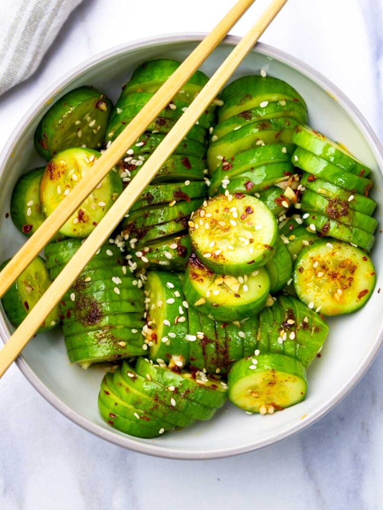 Top-down view of Korean cucumber salad served in a white bowl, drizzled with spicy chili oil dressing and sprinkled with sesame seeds, with chopsticks resting on top.