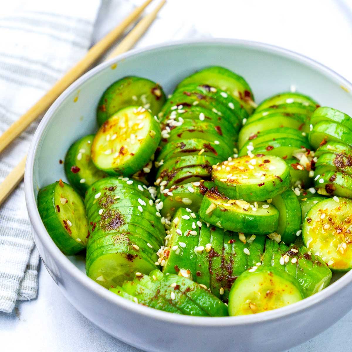Side-angled shot of spicy Korean cucumber salad in a white bowl, showing accordion-style cucumber slices coated in dressing and garnished with sesame seeds.