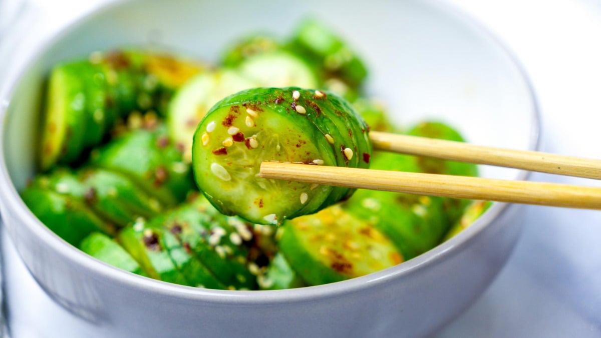 Close-up of chopsticks holding a piece of Korean-style cucumber salad with visible sesame seeds and chili flakes.