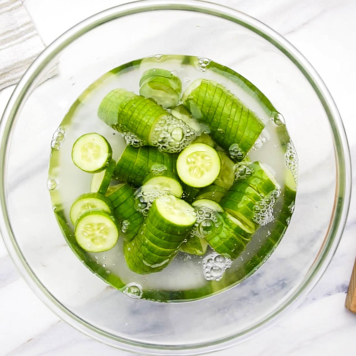 Sliced cucumbers soaking in salted water in a clear bowl to draw out excess moisture.