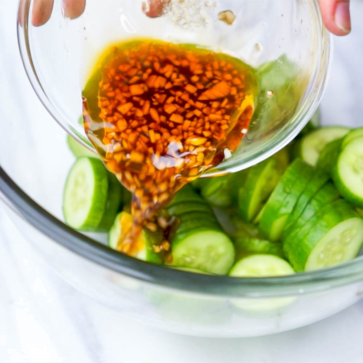 Pouring garlic chili oil dressing over salted cucumber slices in a mixing bowl.